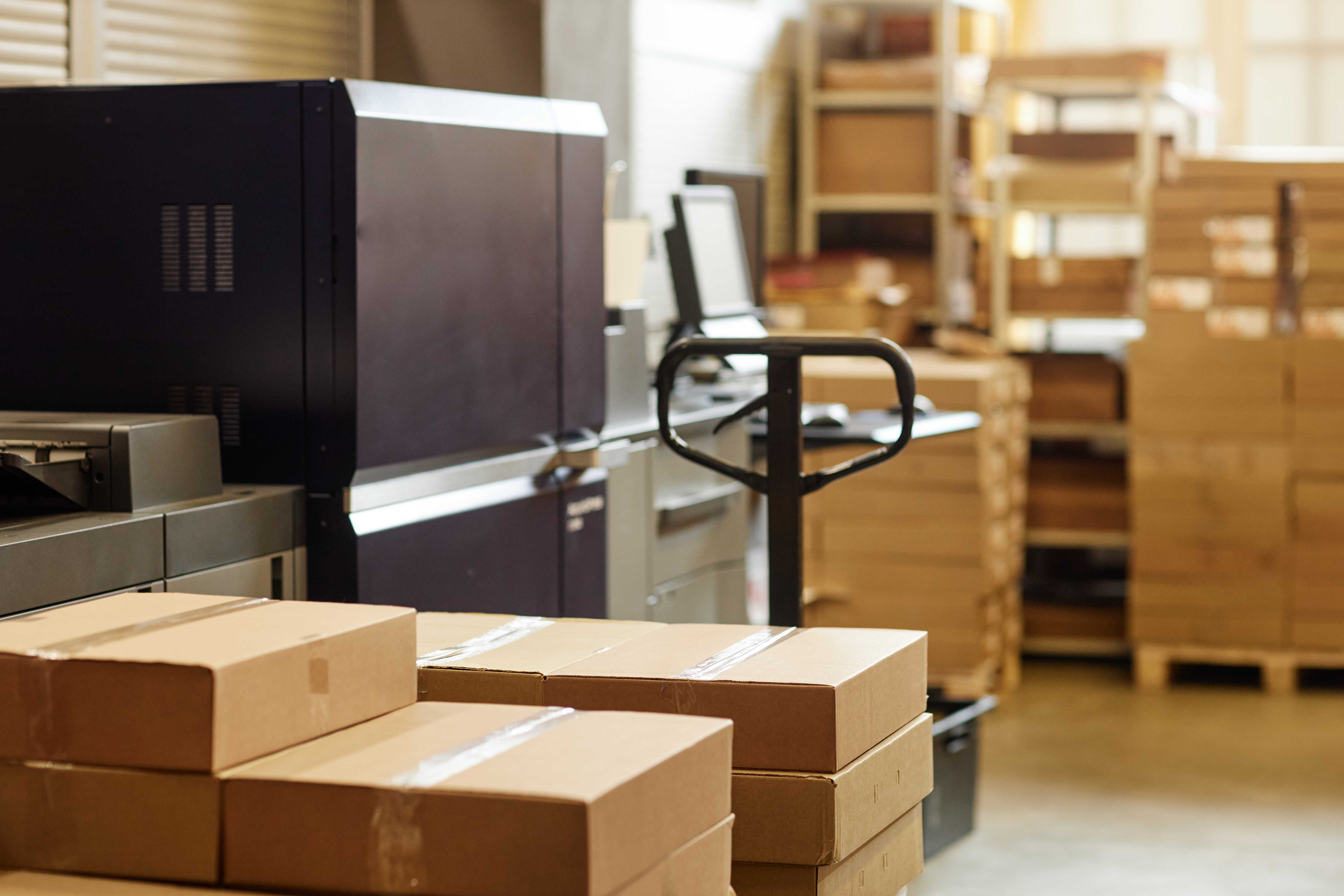 Warehouse fulfillment area with stacked cardboard shipping boxes, industrial printing equipment, and pallets prepared for packaging and distribution.