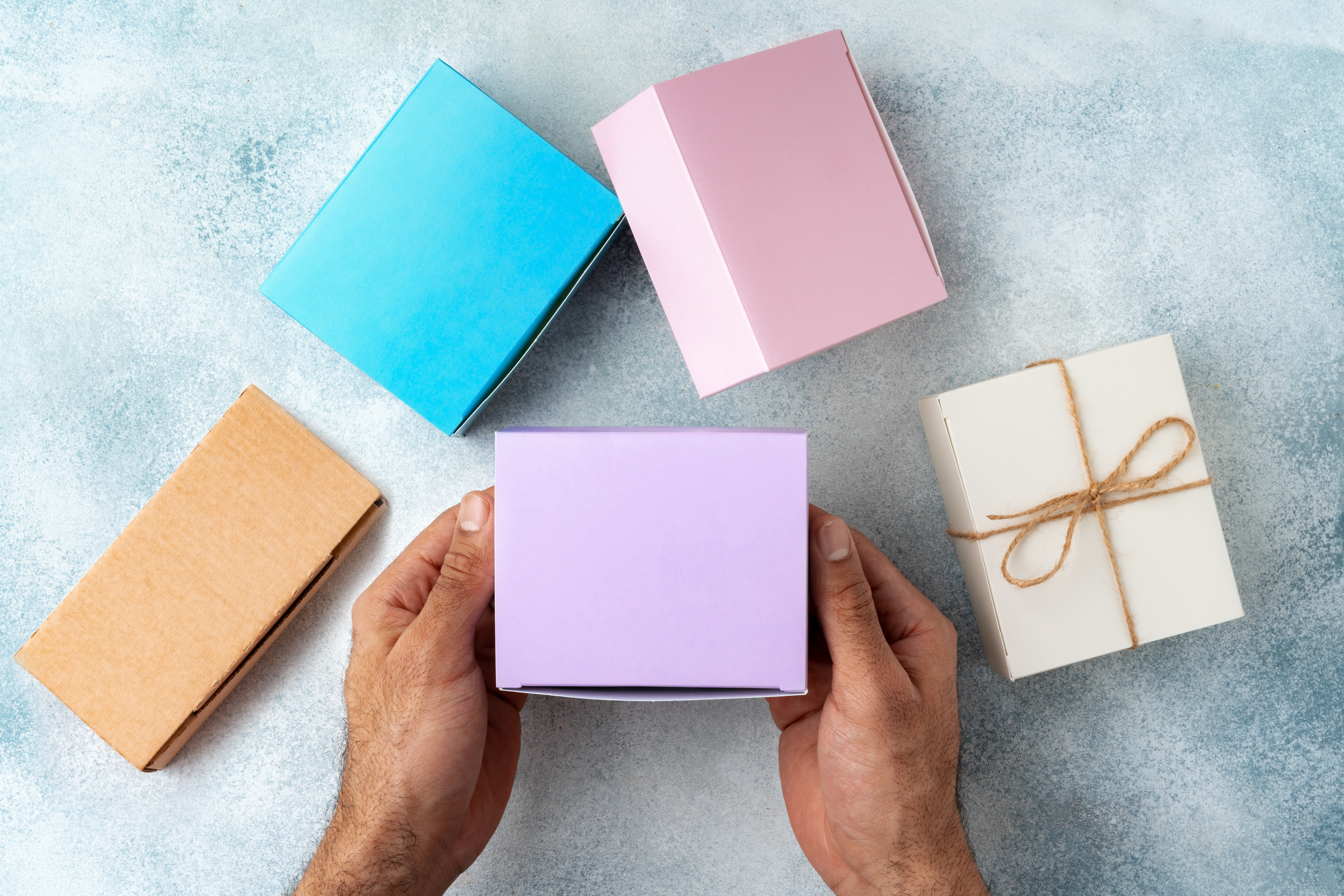 Hands holding a pastel purple gift box surrounded by colorful packaging boxes on a light textured background, showcasing gift wrapping and product packaging options.