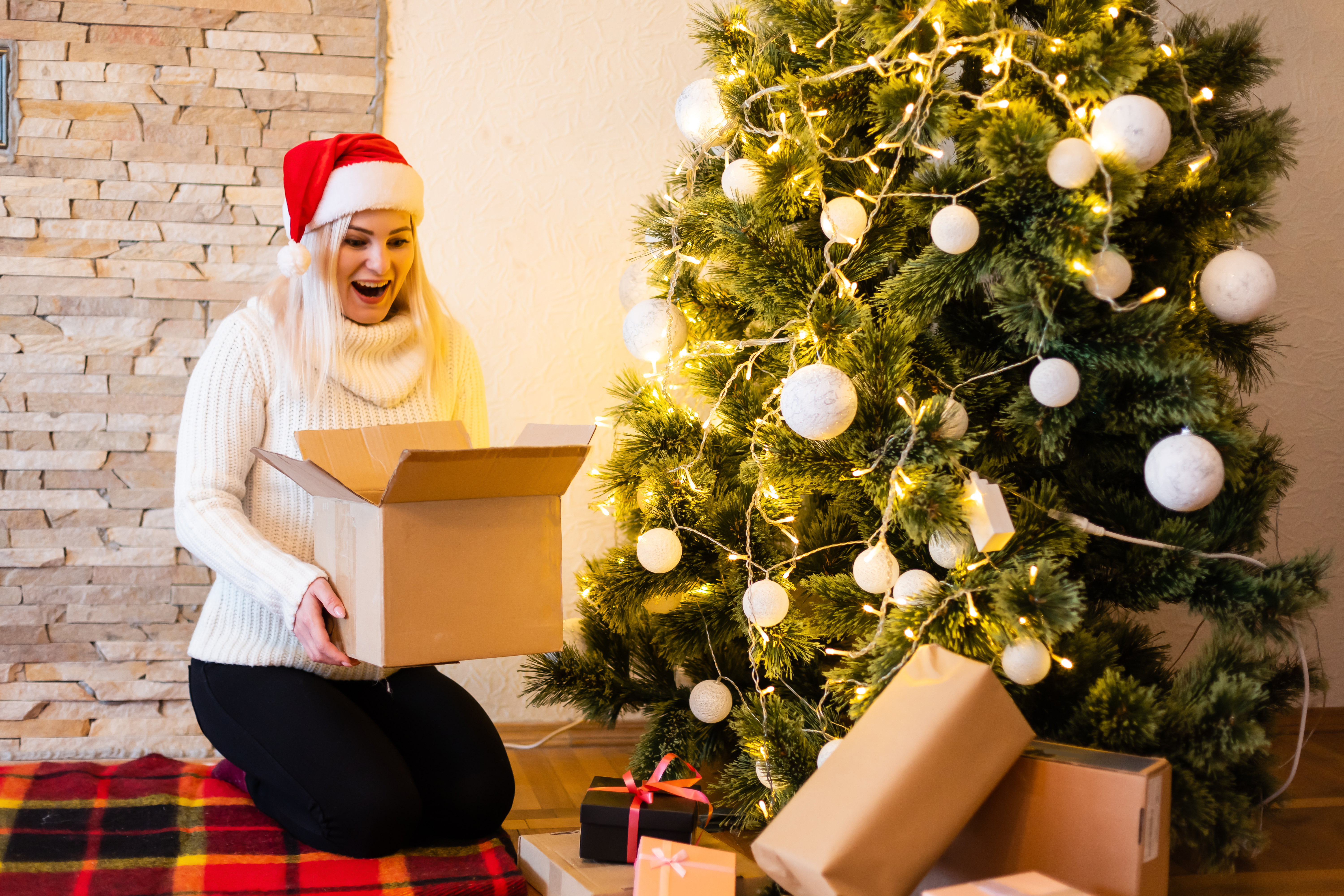 woman sitting by christmas tree smiling at christmas package she is opening 