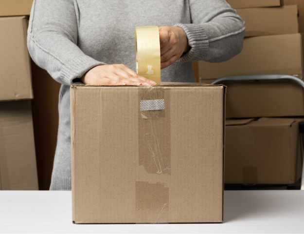 woman taping up a medium cardboard box with stacks of boxes behind her